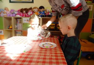 Pani Ewelina nalewa dzbankiem wodę na talerzyk z cukierkami dziewczynki.Chłopiec obserwuje jak rozpuszczają się kolorowe cukierki w wodzie.